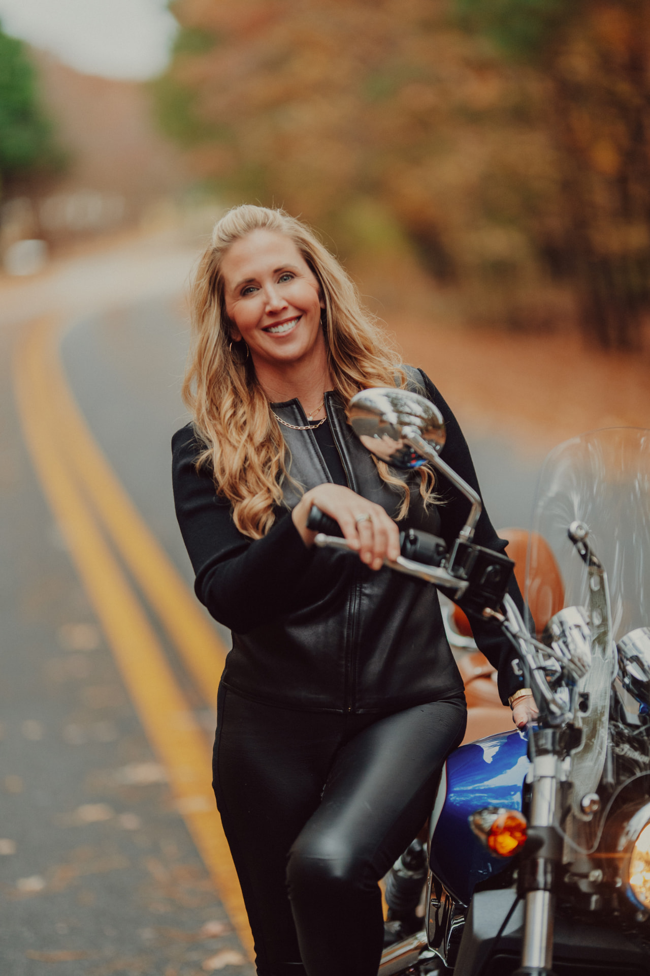 Ashley Davis leaning on her motorcycle on a tree-lined road in autumn